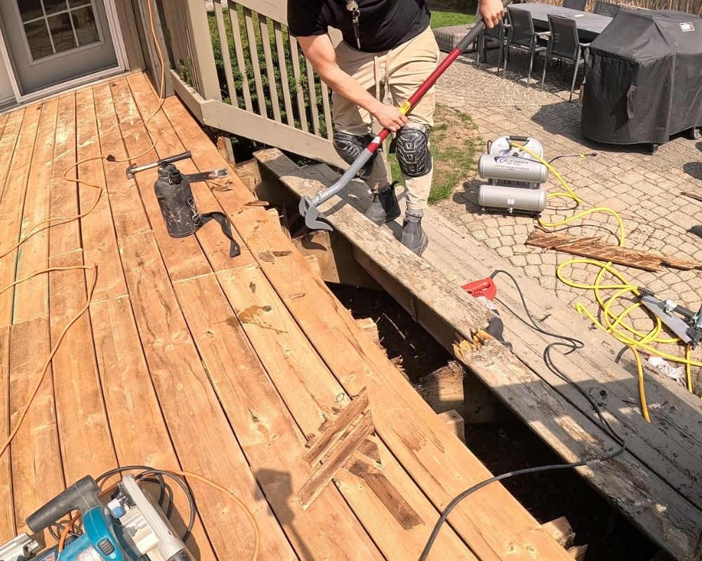 Handyman using a heavy-duty pry bar to carefully remove rotted wood boards during a deck repair project in Oakville.