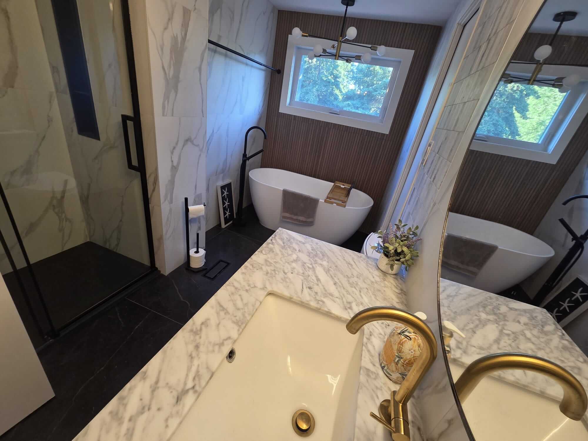 Comprehensive view of a modern master bathroom renovation in Oakville by Bespoke Handyman, featuring a glass shower, freestanding tub, and a marble vanity with brass fixtures, all reflected in a large mirror.