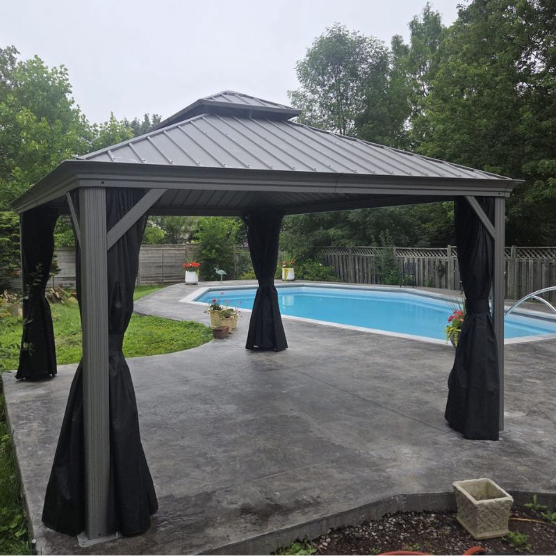 A dark grey metal hardtop gazebo with black privacy curtains and netting sits on a concrete patio next to a backyard swimming pool.